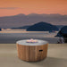 Westminster Teak Agora Fire Table burns with a warm flame on an outdoor terrace overlooking a lake and mountains at sunset