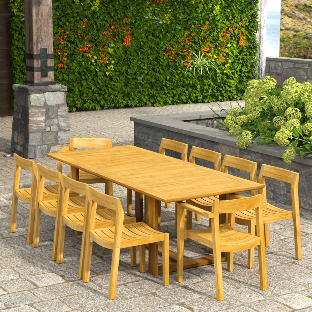 Westminster Teak Horizon 11pc Teak Dining Set is arranged on a patio with a long teak table and matching chairs surrounded by greenery and stone walls