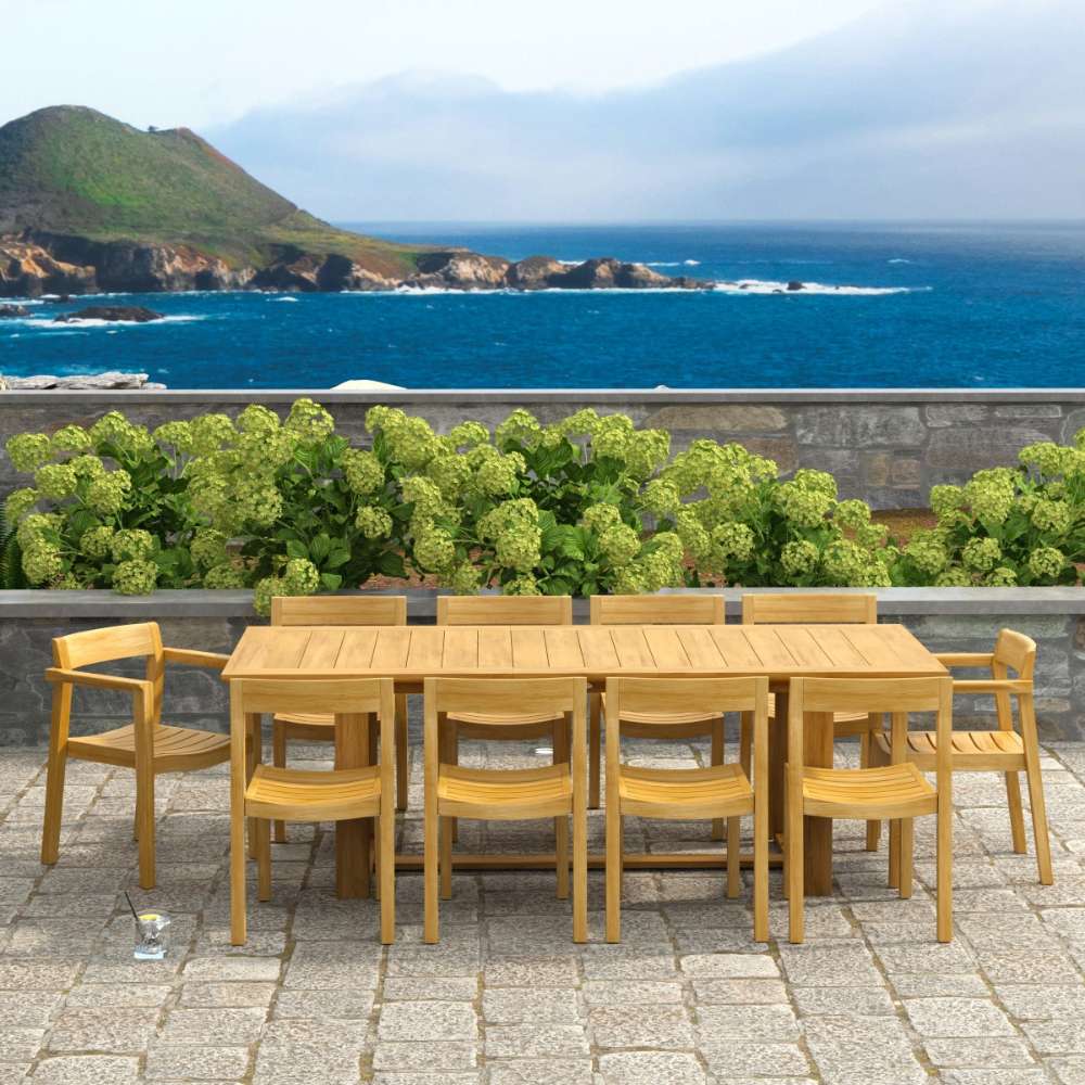 Westminster Teak Horizon 11pc Teak Dining Set is outdoors on a stone patio with the ocean and hills in the background behind the long teak table and matching chairs