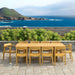 Westminster Teak Horizon 11pc Teak Dining Set is outdoors on a stone patio with the ocean and hills in the background behind the long teak table and matching chairs