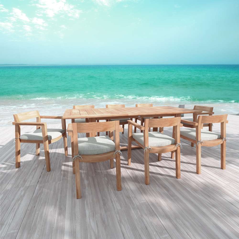 Westminster Teak Horizon 9pcs Teak Dining Set placed on a wooden deck by the ocean, showing the full teak table and chairs in a bright seaside setting
