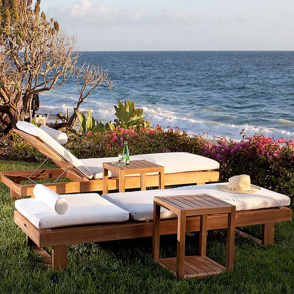 Westminster Teak Horizon Double Chaise and Side Table Set placed on seaside grass with white cushions, two teak side tables, a bottle and glass, and ocean waves behind it