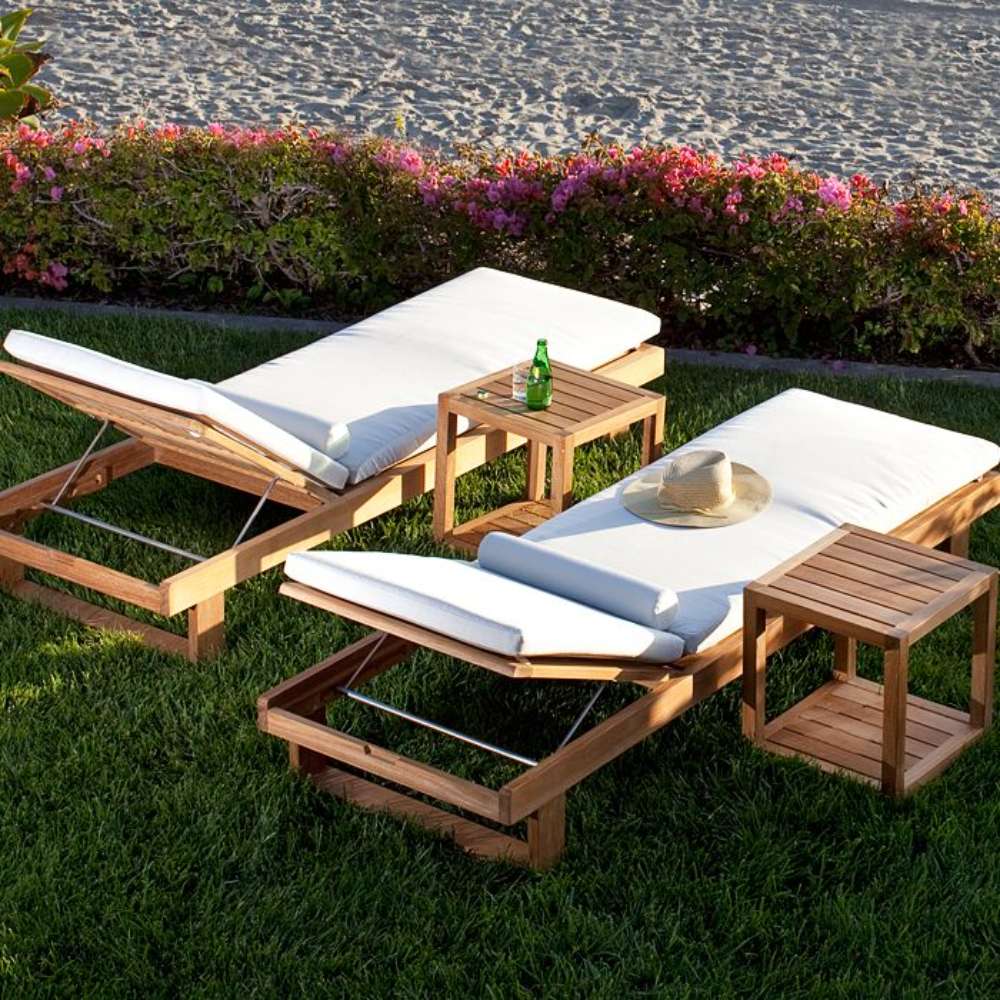 Westminster Teak Horizon Double Chaise and Side Table Set shown on a lawn near the beach with two white-cushioned teak loungers, two side tables, a bottle, and a sun hat
