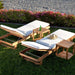 Westminster Teak Horizon Double Chaise and Side Table Set shown on a lawn near the beach with two white-cushioned teak loungers, two side tables, a bottle, and a sun hat