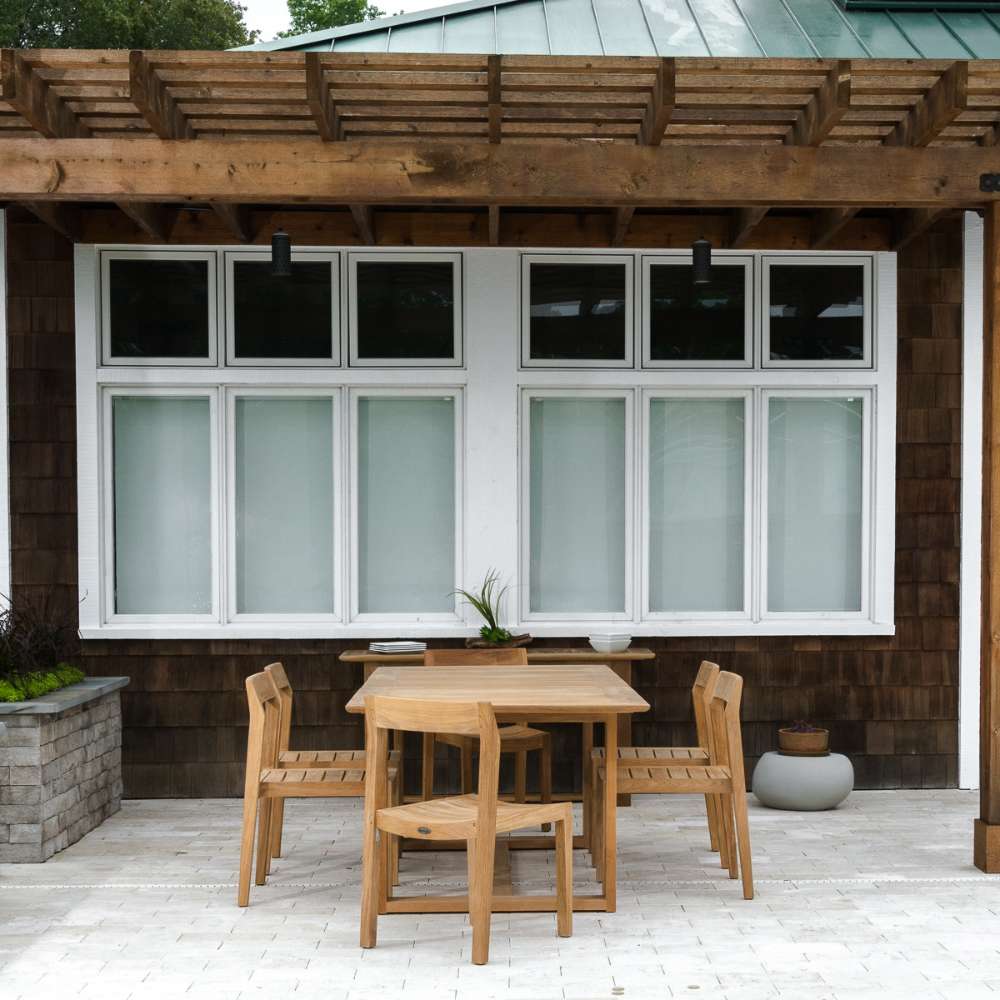 Westminster Teak Horizon Sidechair Dining Set for 6 shows the outdoor teak dining set from farther back under a wooden pergola, centered in front of a house