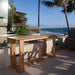 Westminster Teak Horizon Teak Console Table is placed on an outdoor terrace overlooking the ocean, styled with glasses, plates, and a bowl, surrounded by palm trees and greenery