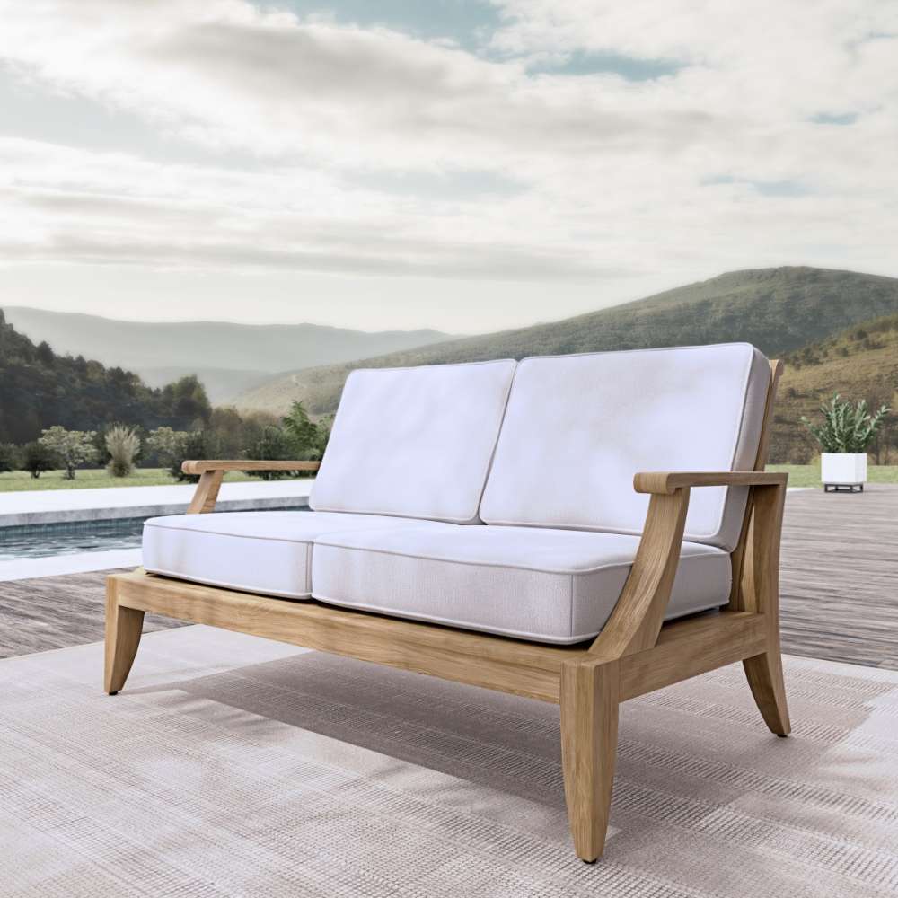 Westminster Teak Laguna Love Seat is placed outdoors on a patio near a pool, showing its natural wood frame and plush cushions in a scenic setting