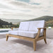 Westminster Teak Laguna Love Seat is placed outdoors on a patio near a pool, showing its natural wood frame and plush cushions in a scenic setting