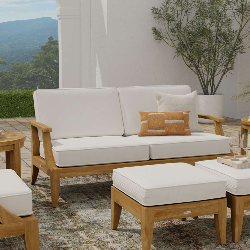 Westminster Teak Laguna Sofa is displayed front-facing on a patio with white cushions, accent pillows, a potted plant, and a scenic outdoor backdrop