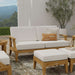 Westminster Teak Laguna Sofa is displayed front-facing on a patio with white cushions, accent pillows, a potted plant, and a scenic outdoor backdrop