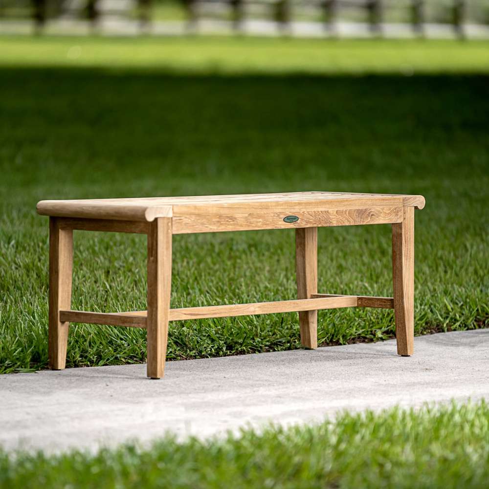 Westminster Teak Laguna Teak Backless Bench is a small, natural wood bench placed on a concrete path with green grass stretching out behind it
