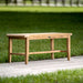 Westminster Teak Laguna Teak Backless Bench is a small, natural wood bench placed on a concrete path with green grass stretching out behind it