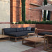 Westminster Teak Maya Chaise complements a modern outdoor patio setup with teak furniture, dark cushions, and a clean architectural backdrop