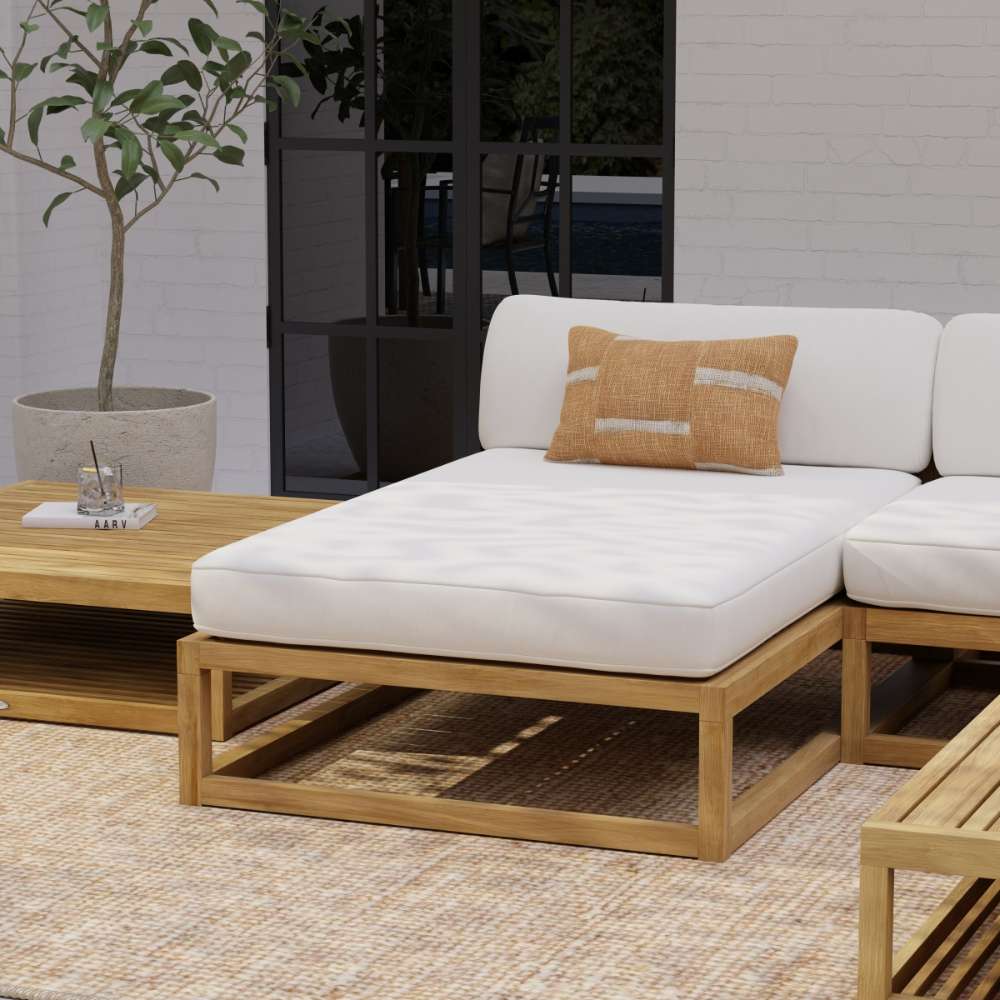 Westminster Teak Maya Chaise features a thick light cushion on a teak frame placed on a patio rug beside a table, potted plant, and glass door