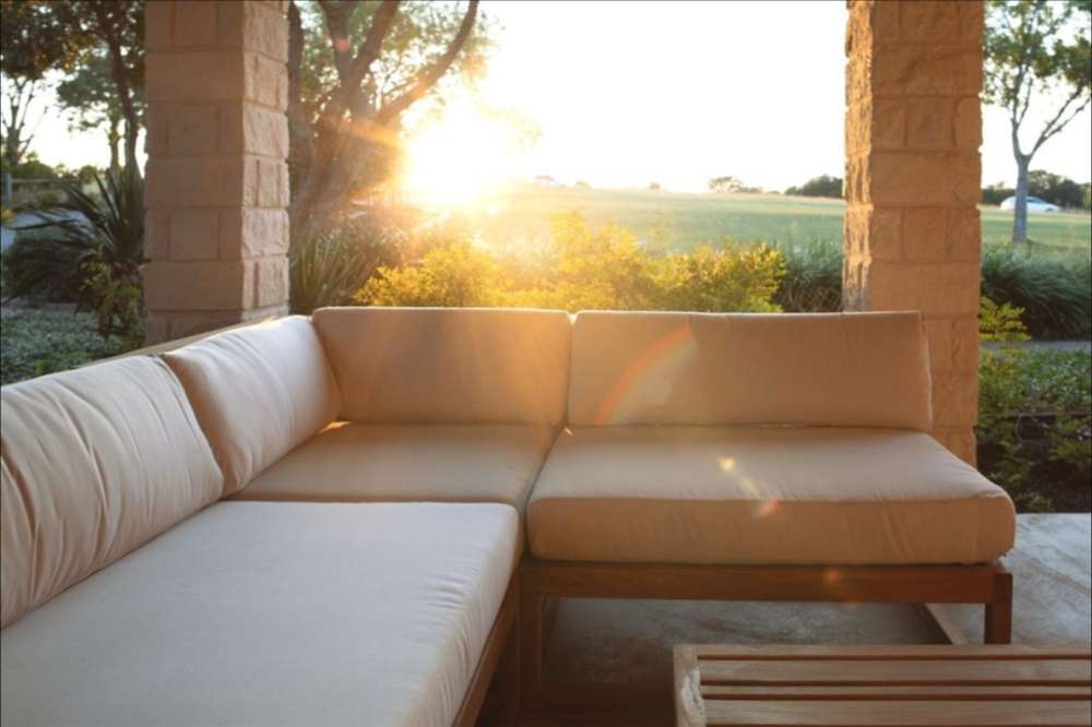 Westminster Teak Maya Corner Sectional is displayed outdoors with cushions, forming a cozy L-shaped seating area facing a sunset over a grassy field