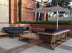 Westminster Teak Maya Left Side Sectional sits on a patio with an umbrella and matching table, set against a brick wall and landscaped plants