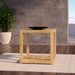 Westminster Teak Maya Teak Side Table is staged on an outdoor rug with a black bowl on top, beside a potted plant and a distant ocean view