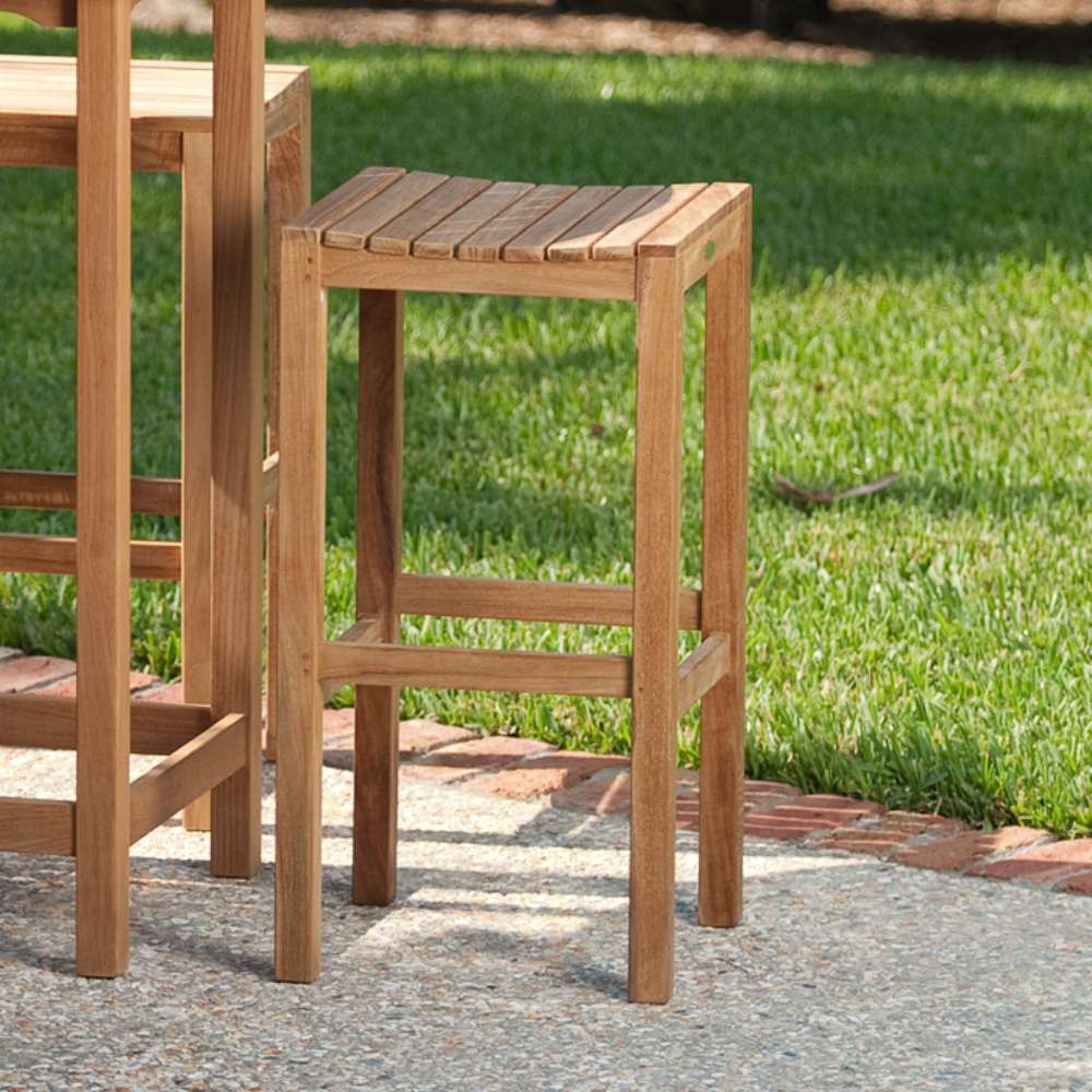 Westminster Teak Somerset Backless Bar Stool is placed outdoors on a patio beside another stool, with green grass in the background