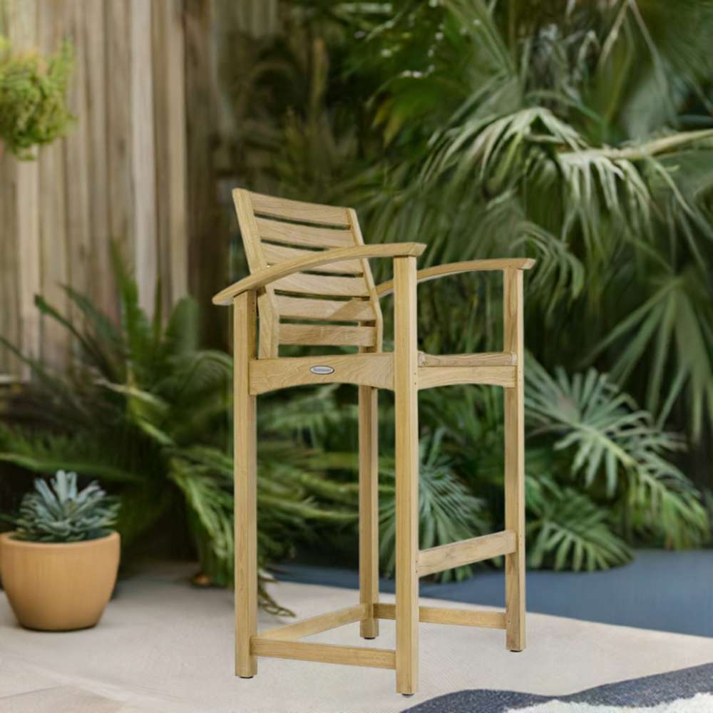Westminster Teak Somerset Teak Bar Stool is placed outdoors on a patio surrounded by lush green plants, showing its use in a garden setting