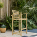 Westminster Teak Somerset Teak Bar Stool is placed outdoors on a patio surrounded by lush green plants, showing its use in a garden setting