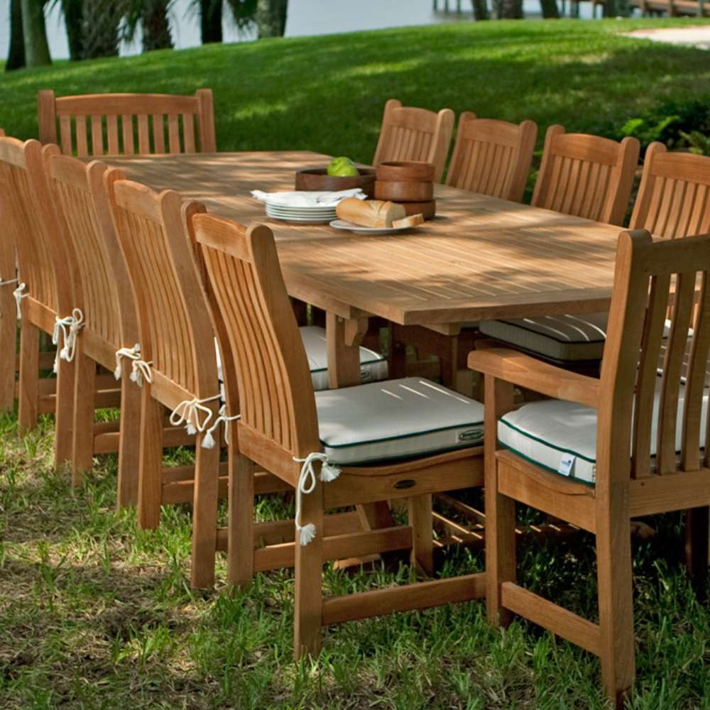 Westminster Teak Veranda Teak Dining Chair lines a large outdoor dining table on grass, complete with cushions, plates, and a scenic lakeside background