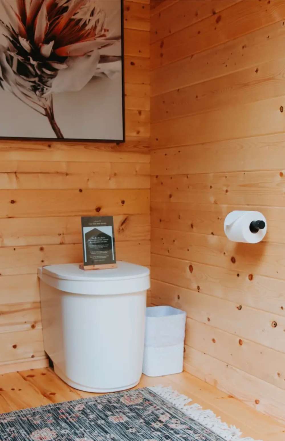 White toilet sits below floral wall art inside Sawmill Structures Loo Loo House Garden Room, paired with a rug and small trash bin