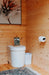 White toilet sits below floral wall art inside Sawmill Structures Loo Loo House Garden Room, paired with a rug and small trash bin