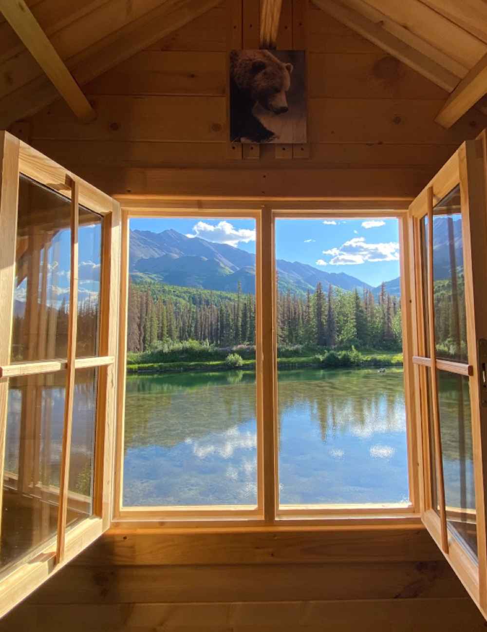 Wide-open windows in the Sawmill Structures The Jumbo Grand Loft 130 SD Garden Room offer a breathtaking view of a calm lake, pine trees, and mountains
