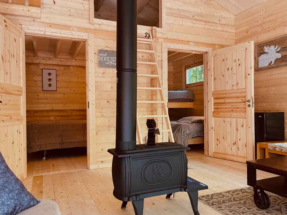 Wood stove stands in the center with two open bedroom doors and a loft ladder inside the Sawmill Structures Edwin Lake House 70MM Garden Room