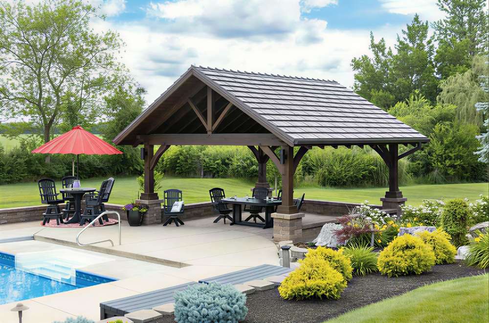 Wooden Brookside Timber Frame Timber Retreat Pavilion covers a spacious patio with tables, chairs, and a nearby pool in a landscaped yard