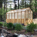 Wooden greenhouse sits among trees and flowers, featuring the 8x12 Outdoor Living Today Western Red Cedar Greenhouse Kit in a natural setting