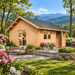 Small wood cabin with a shingle roof, stone patio, and flowers, set against a mountain landscape.
