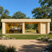 Rectangular light wood cabin with a glass-walled office and open breezeway, situated in a grassy field with trees.