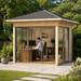 Square wood and glass garden studio with a dark shingle roof, used as a home office with a desk and computer inside.