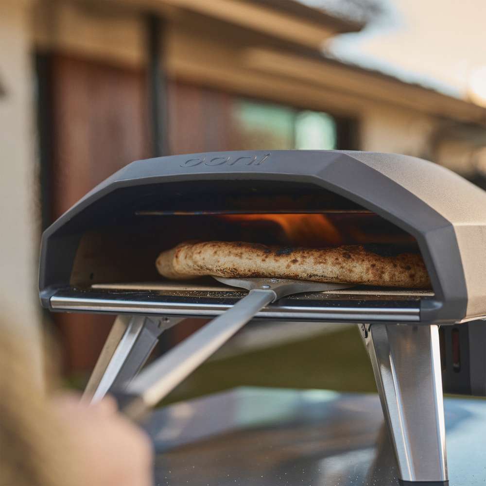 Black Ooni Koda 2 Pizza Oven close view of a pizza on the stone inside the glowing mouth with a peel below.