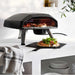 Black Ooni Koda 2 Pizza Oven on a white counter with a pizza cooking inside and a finished pizza on a slate board in front.