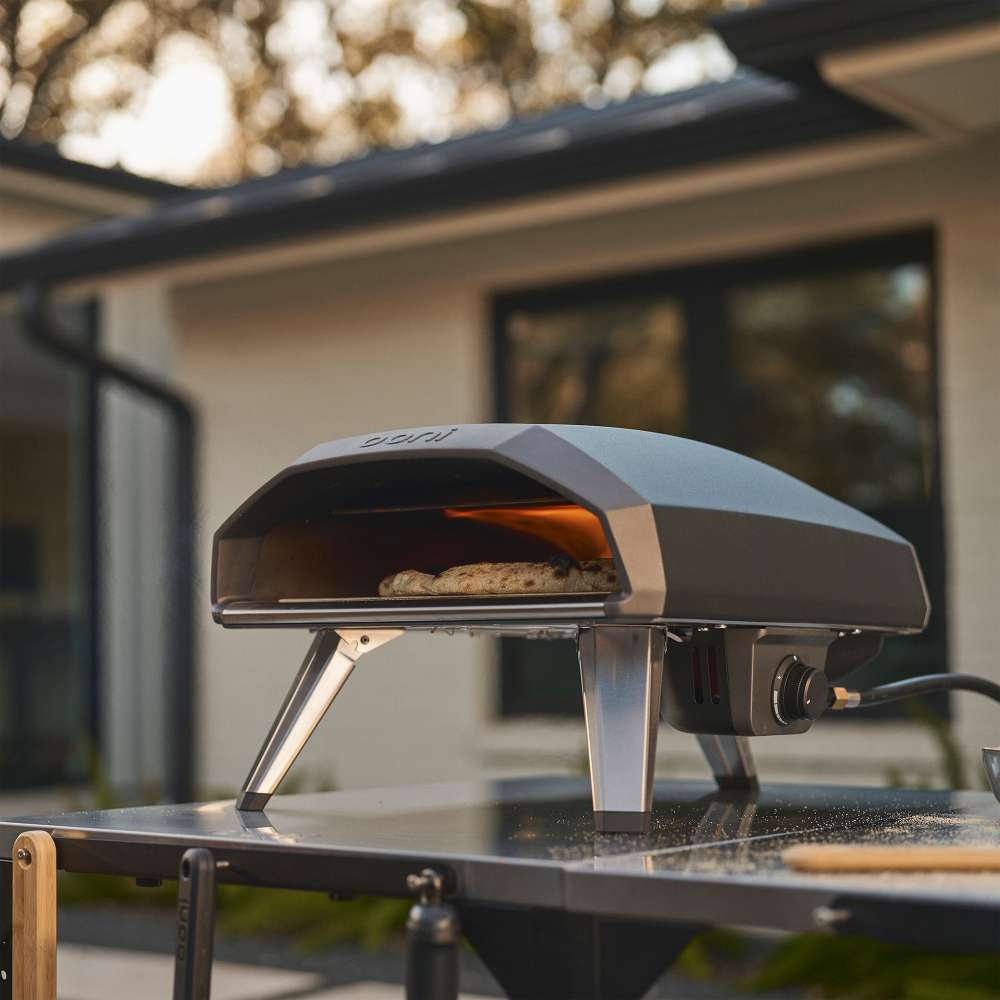Black Ooni Koda 2 Pizza Oven on an outdoor kitchen counter at dusk with a pizza baking in the glowing mouth.