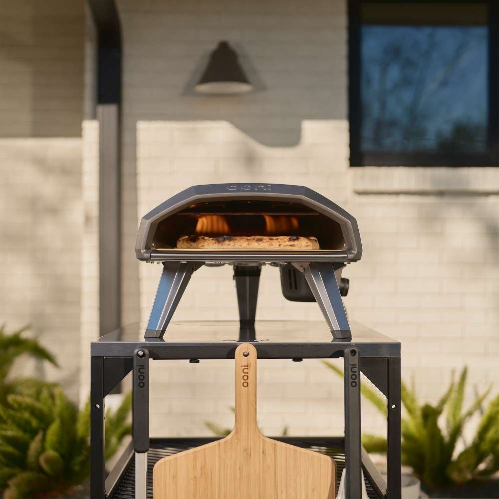 Black Ooni Koda 2 Pizza Oven on an outdoor stand against a white brick wall with a pizza inside and a wooden peel in front.