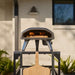 Black Ooni Koda 2 Pizza Oven on an outdoor stand against a white brick wall with a pizza inside and a wooden peel in front.