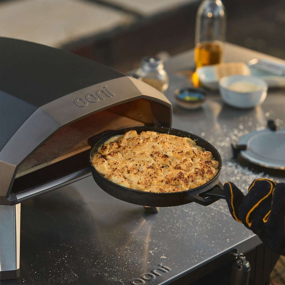 Black Ooni Koda 2 Pizza Oven with a cast iron skillet of bubbling cheese being lifted out onto the oven top.
