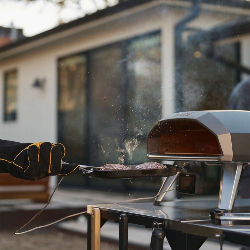 Black Ooni Koda 2 Pizza Oven with a gloved hand sliding a pizza peel toward the mouth outside a modern house.