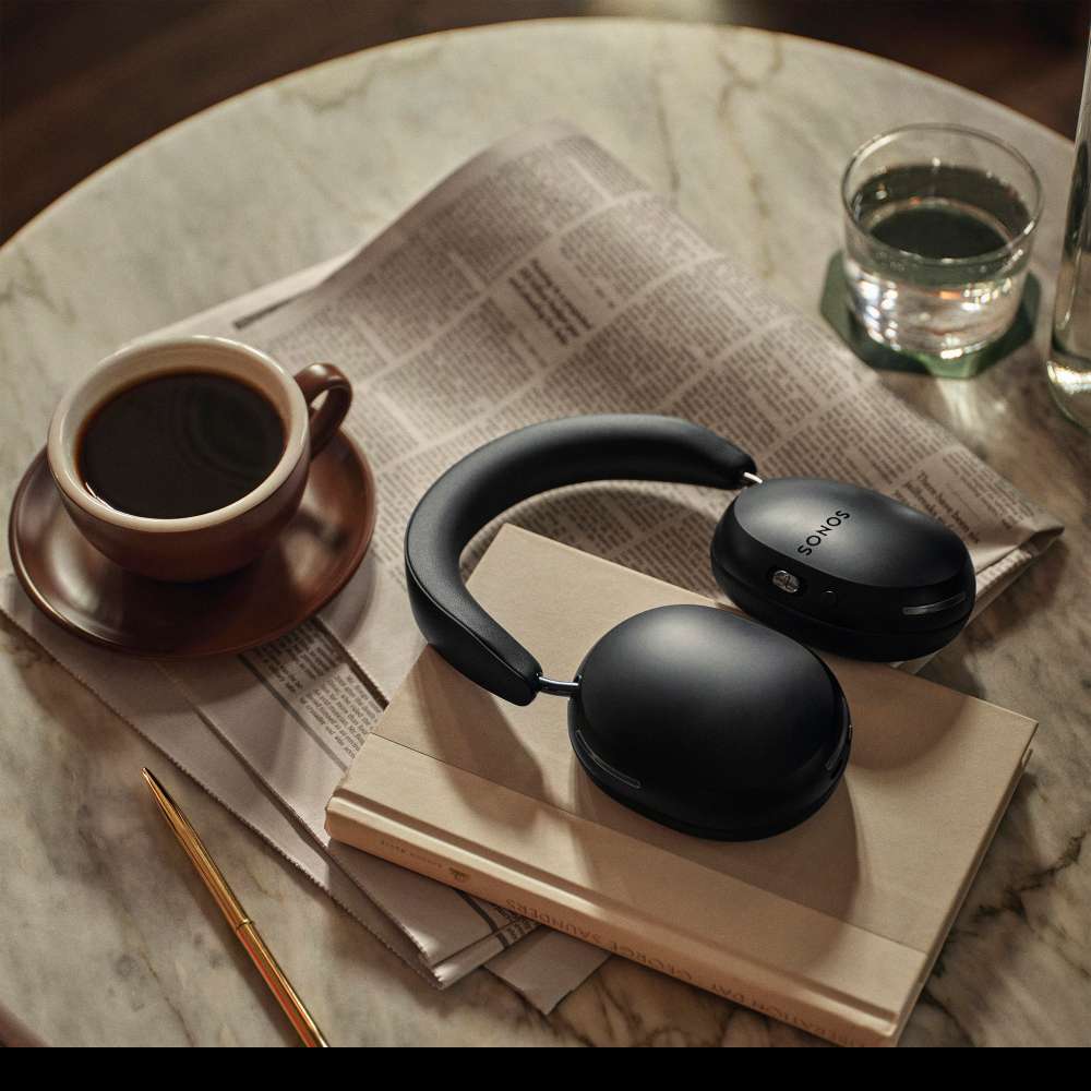 Black Sonos Ace Wireless Headphones with Noise Cancellation resting on a book on a marble cafe table beside a cup of coffee, a glass of water and a newspaper