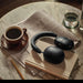 Black Sonos Ace Wireless Headphones with Noise Cancellation resting on a book on a marble cafe table beside a cup of coffee, a glass of water and a newspaper