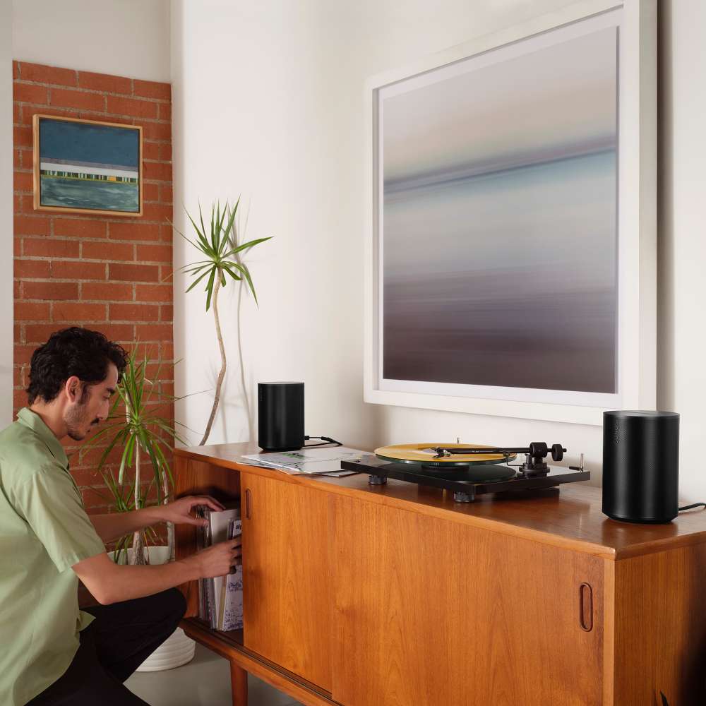 Black Sonos Era 100 Next-Gen Smart Speaker a pair of the speakers on a teak credenza either side of a record player as a man crouches beside it under a large framed print