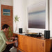 Black Sonos Era 100 Next-Gen Smart Speaker a pair of the speakers on a teak credenza either side of a record player as a man crouches beside it under a large framed print