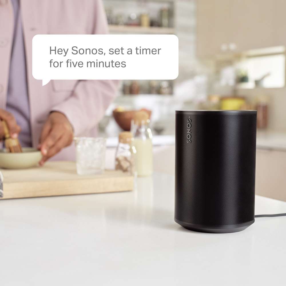 Black Sonos Era 100 Next-Gen Smart Speaker on a white kitchen counter with a speech bubble reading Hey Sonos, set a timer for five minutes while someone cooks in the background