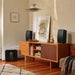 Black Sonos Five Speaker a pair of the speakers standing upright on a teak sideboard either side of a framed picture with a black subwoofer on the floor beside it