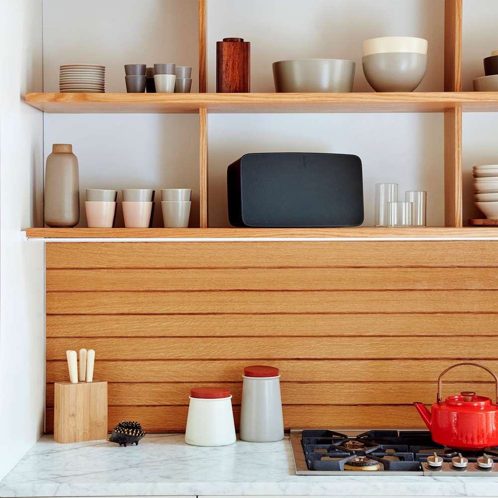 Black Sonos Five Speaker lying horizontally on a kitchen shelf between ceramic cups and bowls above a wooden panelled wall and marble worktop
