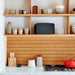 Black Sonos Five Speaker lying horizontally on a kitchen shelf between ceramic cups and bowls above a wooden panelled wall and marble worktop