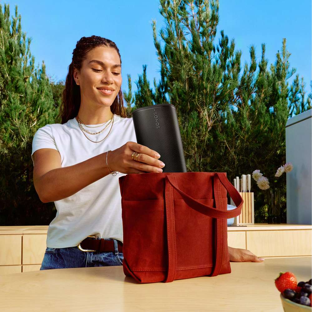Black Sonos Play Portable Bluetooth Speaker being lowered into a red tote bag by a woman standing at an outdoor counter with trees behind her